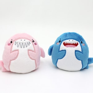 May include: Two plush shark toys, one pink and one blue, with white bellies and embroidered smiling faces.