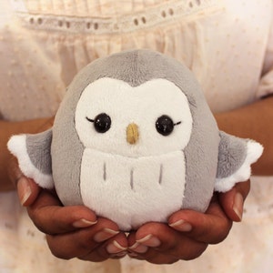 May include: A soft, gray and white plush owl with large black eyes and a small yellow beak. The owl is being held in a person's hands.