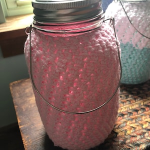 May include: A pink crocheted jar lantern with a silver metal lid and handle. The jar is wrapped in a light pink crochet, and the interior glows with a soft pink light. The lantern sits on a wooden surface.