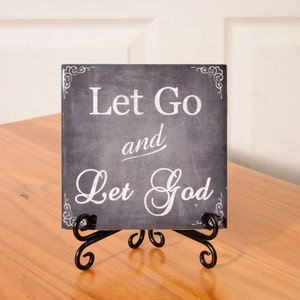 May include: A black chalkboard sign with white lettering that reads "Let Go and Let God". The sign is on a black metal easel.