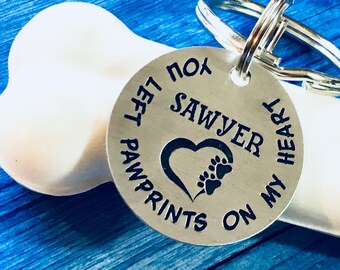 Puede incluir: Una etiqueta de perro de plata con el texto "Left paw prints on my heart, for Sawyer" y un corazón con huellas de patas en su interior.