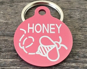 Puede incluir: Etiqueta de identificación redonda para mascotas de color rosa con un anillo plateado. La etiqueta tiene una abeja blanca y la palabra "HONEY" grabada en ella.