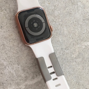 May include: A white Apple Watch with a rose gold case and a grey sport band. The watch face is black and shows a circular design.