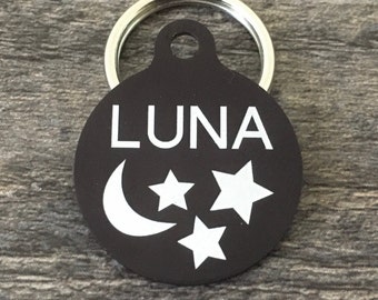 May include: Black round pet tag with a silver ring. The tag has a white crescent moon and three white stars with the name "LUNA" engraved in white letters.