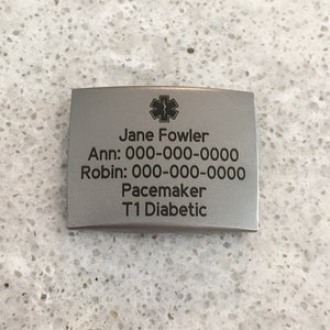 May include: A silver medical alert tag with a star of life symbol, the name Jane Fowler, two phone numbers, and the text "Pacemaker T1 Diabetic".