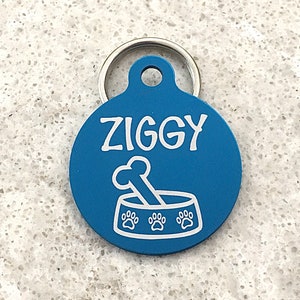 Könnte beinhalten: Runder blauer Hundeanhänger mit einem weißen Knochen und Pfotenabdruck-Design. Der Anhänger trägt die Aufschrift "Ziggy" in weißen Buchstaben.