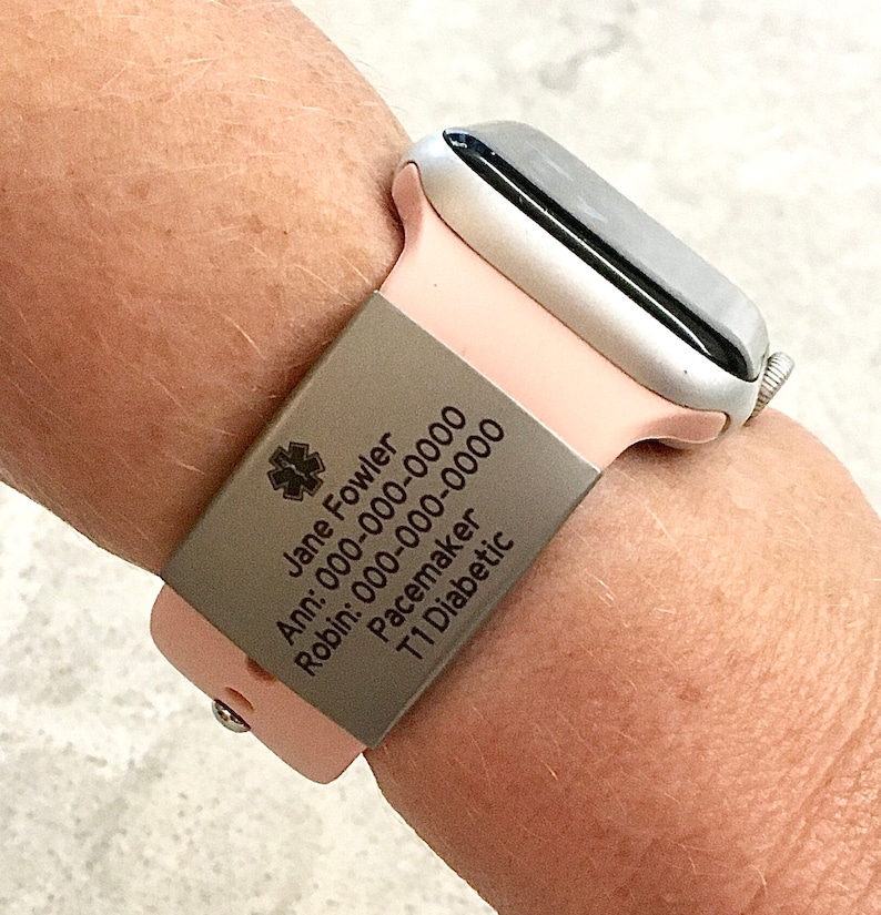 May include: A pink silicone watch band with a silver metal medical alert tag. The tag has the text "Jane Fowler", "Ann: 000-000-0000", "Robin: 000-000-0000", "Pacemaker", and "T1 Diabetic" engraved on it.