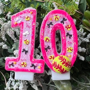 May include: A pair of number candles shaped as 1 and 0, featuring a bright neon pink border and a light pink center filled with iridescent glitter. The candles are decorated with small yellow softball icons and black bow shapes, with a 3D softball attached.