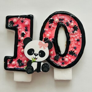 May include: Pink and black number 10 birthday candle with a panda bear holding a bamboo shoot. The candle is decorated with black and white sprinkles.