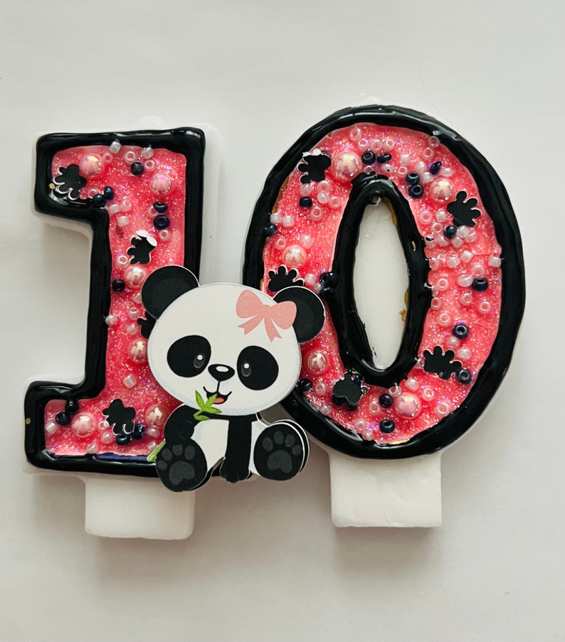 May include: Pink and black number 10 birthday candle with a panda bear holding a bamboo shoot. The candle is decorated with black and white sprinkles.