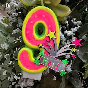 Könnte beinhalten: Eine neonrosa und grüne Geburtstagskerze mit der Zahl neun und Sternen. Die Kerze hat eine grüne Mitte und ist mit kleinen Sternen verziert. Ein kleines Schild mit dem Namen "Heidi" ist angebracht.