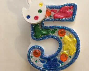 May include: A blue and white birthday candle shaped like the number five. The candle is decorated with colorful paint splatters and glitter. A white paint palette is attached to the top of the candle.