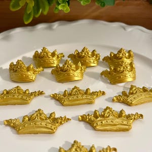May include: A selection of small, gold-coloured crown decorations displayed on a white plate. The crowns feature detailed designs and a metallic finish, indicating they are made of a gold-coloured material. The crowns vary in size and style.