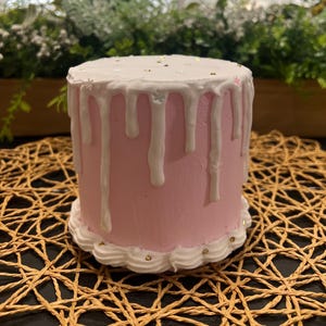 Könnte beinhalten: Ein kleiner, runder rosa Kuchen mit weißer Glasur, die an den Seiten herunterläuft. Die Oberseite ist mit goldenen Streuseln und kleinen weißen schneeförmigen Dekorationen verziert. Der Boden hat einen weißen Zuckergussrand mit goldenen Streuseln.