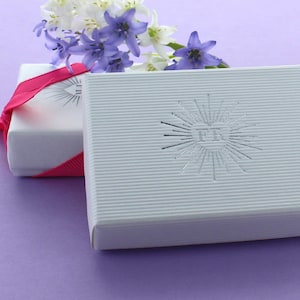 以下が含まれることがあります： ピンクのリボンがかけられた白いギフトボックスが2つあります。右側のボックスには、銀色のエンボス加工が施されたハートの中に「FE」の文字が刻まれています。ボックスは薄い紫色の表面の上に置かれています。