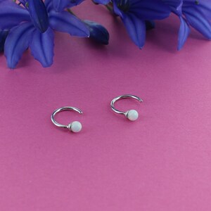May include: A pair of silver hoop earrings with white opal stones. The earrings are on a pink background.