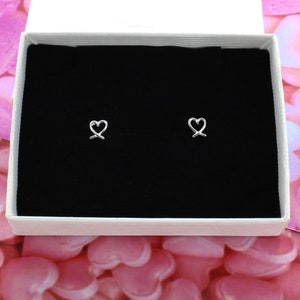 May include: A pair of silver heart-shaped earrings in a white box with a black interior. The earrings are small and delicate.