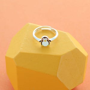 May include: A silver adjustable ring with a penguin design. The penguin has a white body, black wings and head, and a yellow beak and feet. The ring is set against a yellow geometric shape with an orange background.