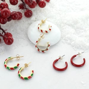 May include: A collection of festive earrings in gold and red, green, and white enamel. The earrings include hoop styles and stud earrings, perfect for holiday or Christmas celebrations. The earrings are displayed on a white surface with red berries.