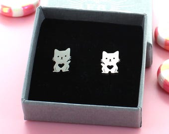 May include: A pair of silver cat earrings with a heart cutout in the centre of each cat. The earrings are in a small grey box with a black interior.