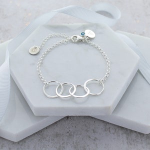 May include: A silver chain bracelet with four interlocking circles. The bracelet has a small silver charm with a blue gemstone and the letter "A" on it.