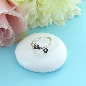 May include: A silver toe ring with a delicate, swirling design. The ring is set on a white stone and has a light blue background.