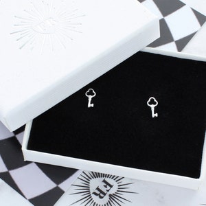 May include: A pair of silver key-shaped earrings in a white box with a black interior. The box has a white starburst design on the lid.