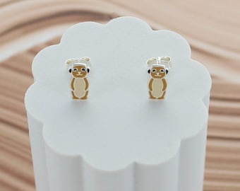 May include: A pair of sterling silver stud earrings featuring a meerkat design. The meerkats have a tan body, white belly, and a silver chef's hat. The earrings are displayed on a white, cloud-shaped stand.