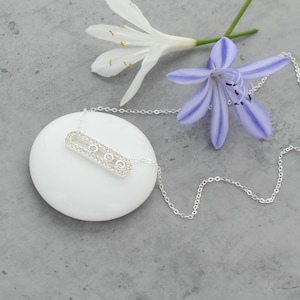 May include: A silver-toned necklace featuring a horizontal rectangular pendant with rounded edges. The pendant is encrusted with small clear stones and contains three circular bezel-set stones arranged in a line along the centre.