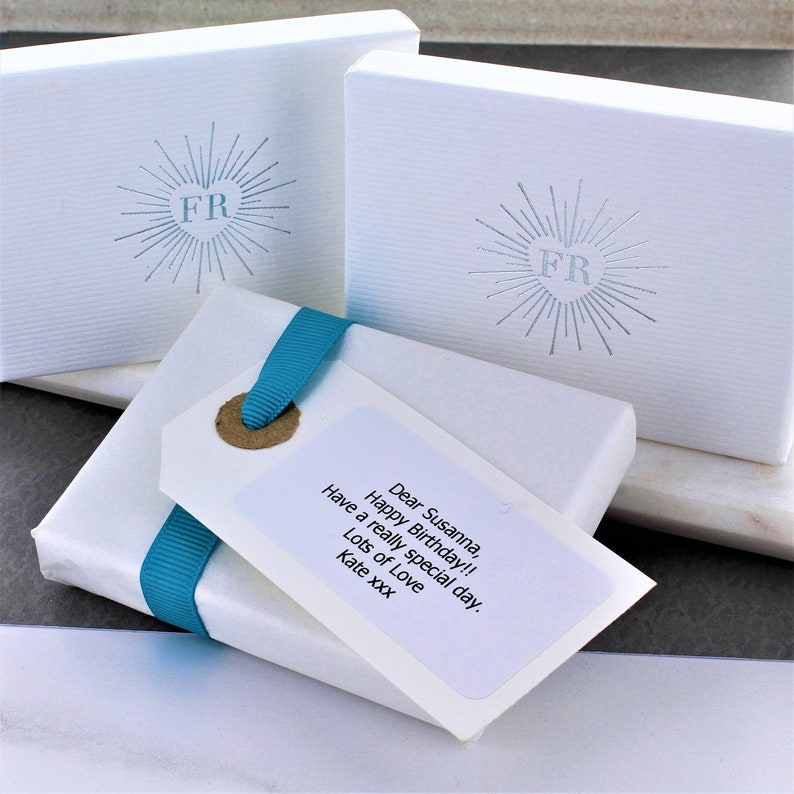 Puede incluir: Dos cajas de regalo blancas con una cinta azul y una tarjeta escrita a mano que dice "Querida Susanna, &iexcl;Feliz cumplea&ntilde;os! Que tengas un d&iacute;a realmente especial. Mucho amor Kate xxx". Las cajas tienen un logotipo plateado en relieve con las letras "FR" dentro de un coraz&oacute;n.