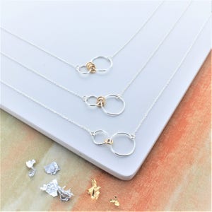 May include: Three silver necklaces with gold circle accents. The necklaces are layered on a white surface. The necklaces feature two interlocking circles, one larger than the other.