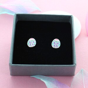 May include: A pair of silver stud earrings featuring a pastel pink, green, and purple enamel design of an Easter egg with a bow. The earrings are presented in a grey box.