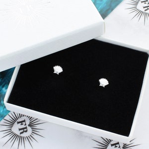 May include: A pair of silver shell stud earrings in a black velvet jewellery box with a white lid. The box has a white logo with the letters "FR" in a circle.