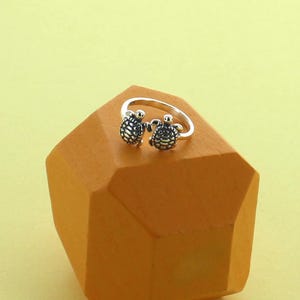 May include: A silver ring with two turtle motifs. The turtles are intricately detailed and sit next to each other on the ring's band. The ring is presented on an orange-brown geometric block against a yellow-green backdrop.