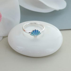 Könnte beinhalten: Ein silberner Ring mit einem kleinen, fächerförmigen Muschel-Anhänger. Die Muschel ist mit einem Farbverlauf aus blauem und türkisfarbenem Emaille verziert, der von einem dunkleren Ton an der Basis zu einem helleren Blau am Rand übergeht.
