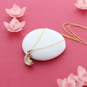 May include: A gold necklace with a small, detailed pasta pendant. The necklace is on a white surface with pink flowers in the background.