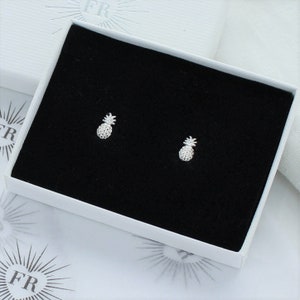 May include: A pair of silver pineapple stud earrings in a black velvet jewelry box. The earrings are small and delicate, perfect for adding a touch of whimsy to any outfit.