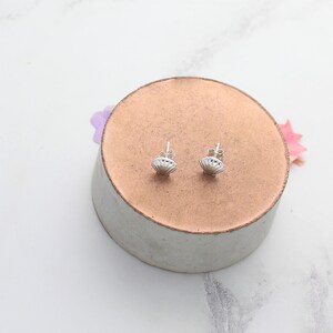 May include: A pair of silver shell-shaped stud earrings on a light brown circular surface.