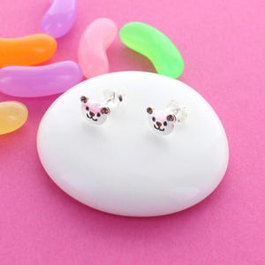 以下が含まれることがあります： ピンクと白のエナメルでクマのデザインが施されたシルバーのスタッドピアスのペア。ピアスは白い表面の上に置かれています。