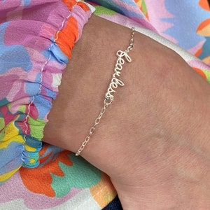 May include: A silver bracelet with a delicate chain and the word "Fearless" written in cursive.