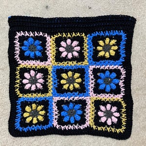 May include: A black crocheted square with nine smaller squares inside. The smaller squares are in alternating colors of pink, blue, and yellow with a black center.