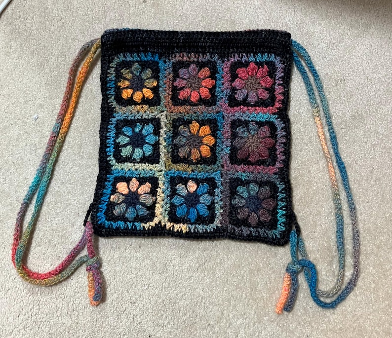 May include: A crocheted drawstring bag with a black background and colorful floral patterns. The bag has two long, colorful straps.