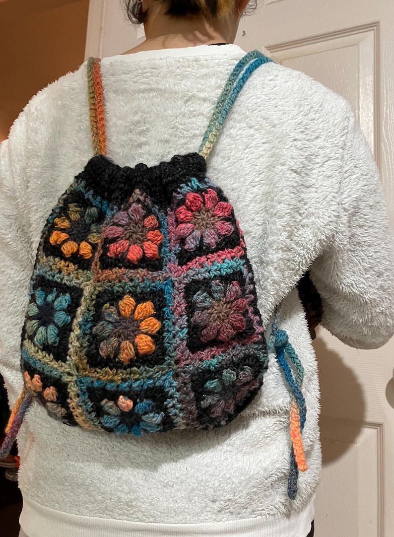 May include: A colorful crocheted backpack with a drawstring closure. The backpack is made of multiple squares with different floral patterns in shades of pink, orange, blue, green, and brown.