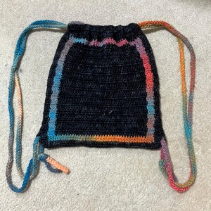 May include: A black crocheted drawstring bag with a multi-colored border. The bag has two long drawstrings that are also multi-colored.
