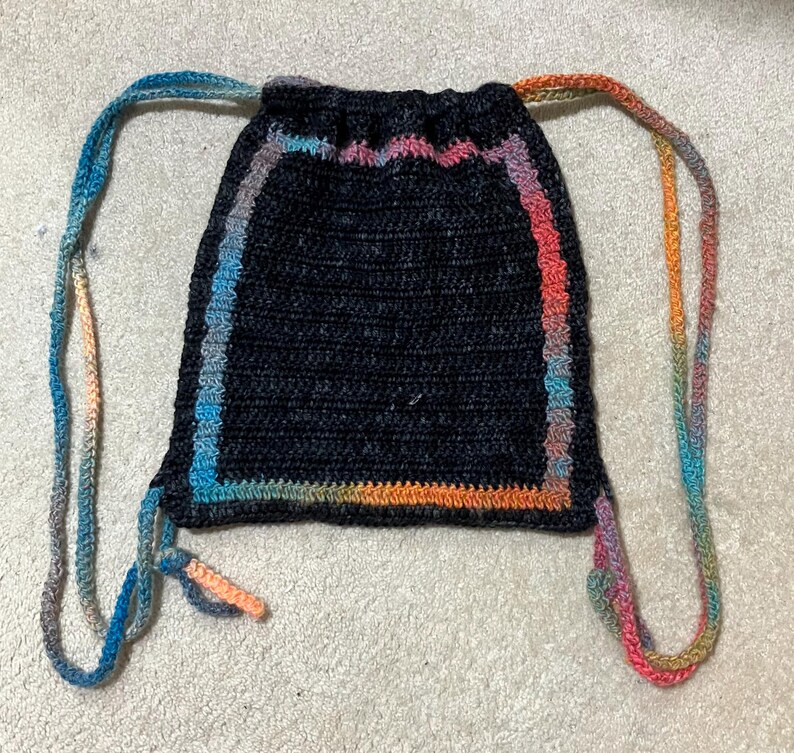 May include: A black crocheted drawstring bag with a multi-colored border. The bag has two long drawstrings that are also multi-colored.