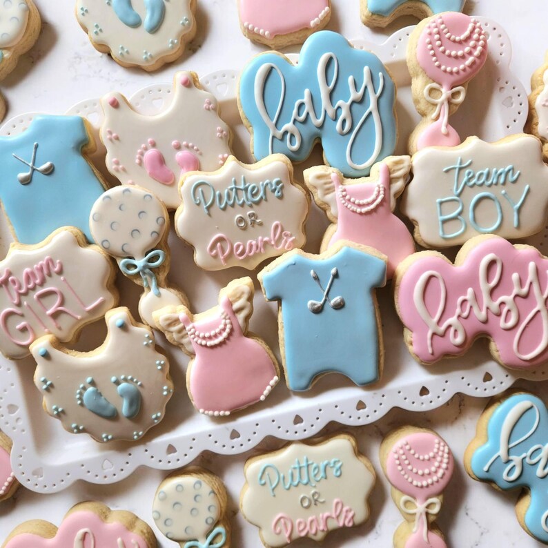 May include: Assortment of baby shower cookies in various shapes and colors. Cookies include baby bibs, dresses, rattles, and shirts, decorated with pink and blue icing. Some cookies have the words "baby", "Team Boy", "Team Girl", and "Putters or Pearls".