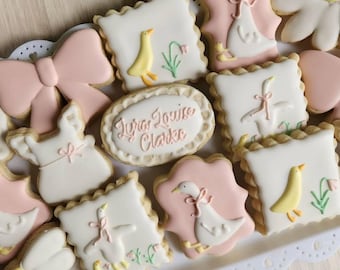 May include: A collection of decorated sugar cookies in a variety of shapes, including squares, ovals, and flowers. The cookies are decorated with pink, white, and yellow icing, featuring designs of geese, bows, and the text "Lyra Louise Clarke".