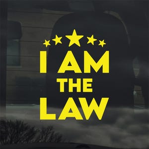 May include: Yellow vinyl decal with the text "I AM THE LAW" and a row of stars above the word "AM". The decal is designed to be applied to a car window or similar surface. The text is in a bold, sans-serif font.