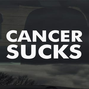 Puede incluir: Adhesivo para ventana de coche con el texto blanco «CANCER SUCKS» sobre un fondo oscuro. La imagen sugiere una calcomanía, con una vista borrosa a través del cristal. El mensaje es una declaración de apoyo.