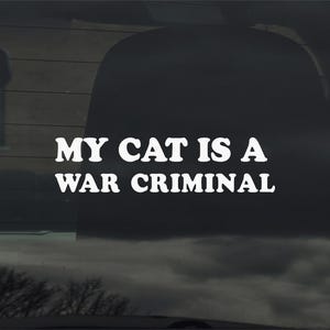 May include: White vinyl decal with the text "MY CAT IS A WAR CRIMINAL" in a bold, sans-serif font. The decal is applied to a car window, with a dark interior visible behind it. The background includes a cloudy sky and tree branches.